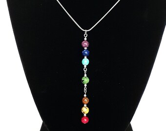 Può includere: Una collana a catena color argento con un pendente verticale composto da sette piccole perline sferiche multicolori nelle tonalità viola, blu, turchese, verde, marrone, giallo e rosso, separate da minuscoli distanziatori in metallo argentato.