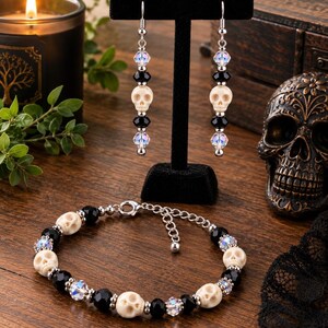 May include: A pair of drop earrings and a matching bracelet featuring off-white skull beads, faceted black beads, and iridescent clear crystal beads. The jewelry set includes silver-toned metal components and a lobster claw clasp on the bracelet.