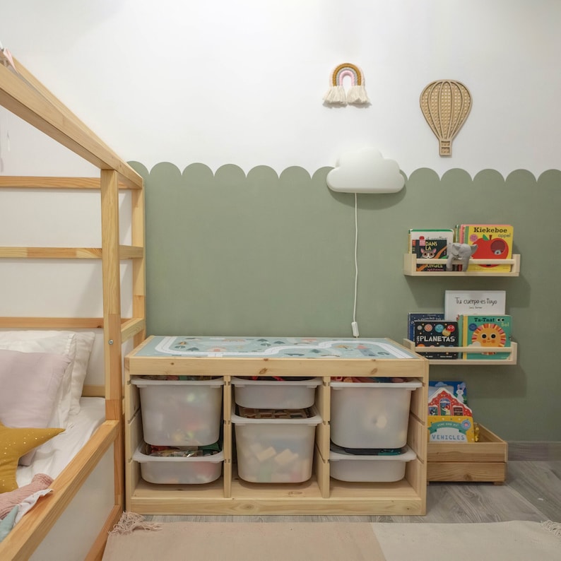 K&ouml;nnte beinhalten: Ein Holzspielzeugregal mit wei&szlig;en Plastik-Aufbewahrungsbeh&auml;ltern und einer Holzplatte. Das Regal ist gef&uuml;llt mit Kinderb&uuml;chern und Spielzeug. Das Regal steht an einer gr&uuml;nen Wand mit einer wei&szlig;en, wolkenf&ouml;rmigen Nachtlampe.