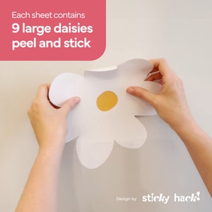 May include: A large white daisy peel and stick decal with a yellow centre is held up against a white wall. The text in the upper left corner reads, "Each sheet contains 9 large daisies peel and stick."