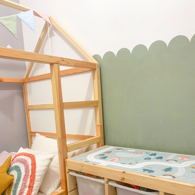 K&ouml;nnte beinhalten: Ein Holzbettgestell mit einem wei&szlig;en Kopfteil und einer gr&uuml;nen Wand mit einer gewellten Bord&uuml;re. Das Bett ist mit einer wei&szlig;en und grauen gemusterten Decke und einem regenbogenfarbenen Kissen bezogen. Im Vordergrund steht ein Holztisch mit einer Spielmatte, die eine Stra&szlig;e und B&auml;ume zeigt.