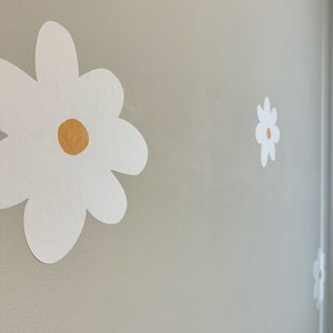 May include: White daisy flower wall decals with a yellow centre are affixed to a light grey wall. The decals are of varying sizes and are arranged in a scattered pattern, creating a whimsical floral design.