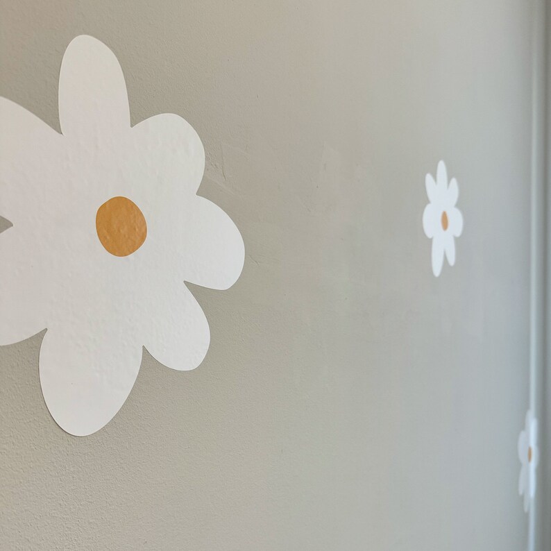May include: White daisy flower wall decals with a yellow centre are affixed to a light grey wall. The decals are of varying sizes and are arranged in a scattered pattern, creating a whimsical floral design.