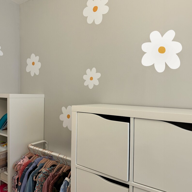 May include: A room featuring white daisy wall stickers with yellow centres on a light grey wall. White storage units and a clothing rail with colourful children's clothing are also visible.