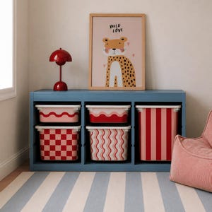 Op de afbeelding: Een blauwe opbergkast met vijf rechthoekige bakken met rode en roze patronen, waaronder strepen, golven en ruiten. Bovenop staan een rode paddenstoelvormige lamp en een ingelijste print met een luipaardillustratie en de tekst Wild Love.