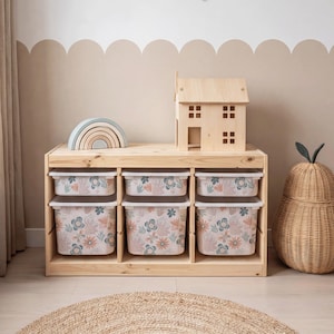 Puede incluir: Un mueble de almacenamiento de madera con seis contenedores con estampado floral. Una casa de madera y un arcoíris están encima. Una cesta trenzada en forma de pera y una alfombra redonda de yute completan la escena. Los contenedores están decorados con flores rosas, azules y beige.