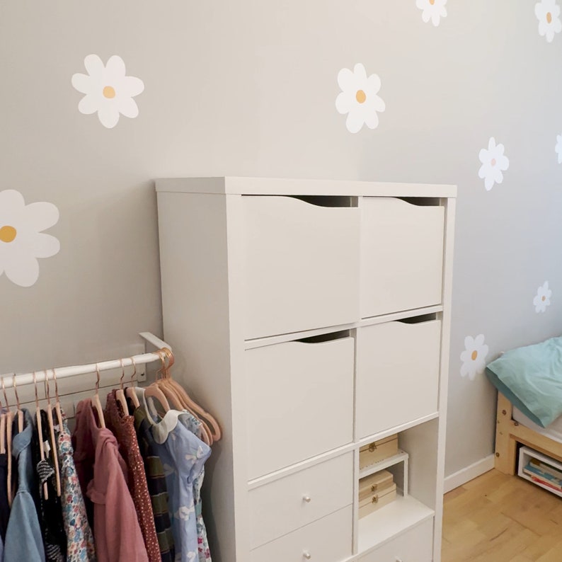 May include: A white storage unit with drawers and cubbies is set against a light grey wall decorated with daisy decals. A clothing rail with pink hangers and children's clothes is in the foreground. A bed with a blue cushion is visible.