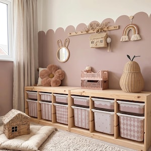 Puede incluir: Una habitación infantil con una combinación de colores rosa y blanco. Unidades de almacenamiento de madera con contenedores estampados. Los elementos decorativos incluyen una almohada de flores, un espejo con forma de conejo, una casa tejida y una cesta en forma de pera. También se ve un adorno de pared de arcoíris y un letrero que dice "magic".