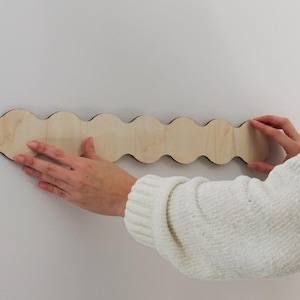 Peut inclure: Un élément de décoration murale en bois clair avec un motif ondulé. La pièce est maintenue contre un mur blanc. Les bords du décor sont sombres, créant un contraste avec le bois clair. Le décor mesure environ 76 cm de long.
