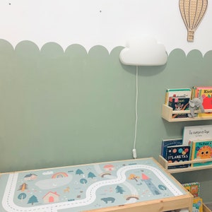 K&ouml;nnte beinhalten: Eine hellgr&uuml;ne Wand mit einem wei&szlig;en gewellten Rand und einem wei&szlig;en, wolkenf&ouml;rmigen Nachtlicht. Ein Holzregal mit B&uuml;chern in Spanisch, Englisch und Franz&ouml;sisch. Eine Spielmatte mit einer Stra&szlig;e und B&auml;umen auf einem Holztisch.