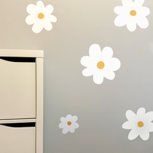 May include: White daisy flower wall decals with yellow centres are displayed on a grey wall. A white cabinet is visible on the left side of the image. The daisies are of varying sizes and are evenly spaced.