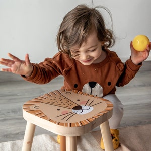 May include: A wooden children's stool with a lion face design. The square seat features a tan lion's face with a white muzzle, whiskers, and a brown mane. The stool has four wooden legs. The child is wearing a brown sweater with a bear face.