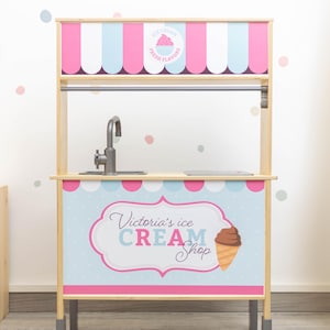 May include: A wooden ice cream stand playset with a pink and blue striped awning. The stand has a sink, a counter, and a sign that says "Victoria's Ice Cream Shop".