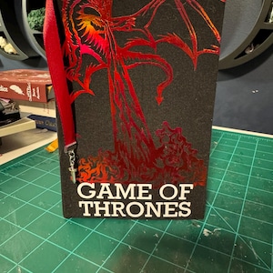 Puede incluir: Un libro de tapa dura negro con el título "GAME OF THRONES" en letras blancas. La portada tiene un diseño de dragón rojo. Una cinta roja de marcador con un dije de espada está adjunta.
