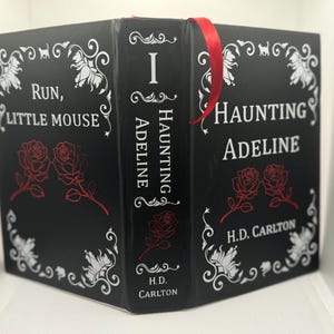 Puede incluir: Un libro negro con el título "Haunting Adeline" y "Run, Little Mouse" en texto blanco. El libro presenta ilustraciones de rosas rojas y un marcapáginas de cinta roja. El nombre del autor, H.D. Carlton, también es visible.
