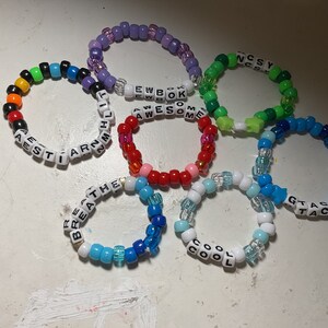 May include: A collection of colourful beaded bracelets with different words spelled out on them, including "AESTI", "ARM", "COOL", "GTAC", "BREATHE", "EWBOKAWSOME", and "NCSY".