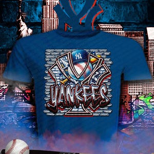 May include: Blue t-shirt featuring a baseball design with the text "Yankees" in a graffiti style. The design includes a baseball, a baseball cap, and the team's logo. A baseball and bat are in the foreground.