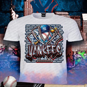 May include: White t-shirt featuring a graphic design of the New York Yankees logo, baseball, and bat. The design incorporates the word "Yankees" in a graffiti-style font, set against a brick wall background. The shirt has a crew neck.