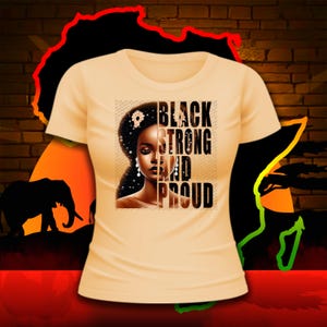 Puede incluir: Una camiseta de color melocot&oacute;n claro con un dise&ntilde;o gr&aacute;fico en blanco y negro. El gr&aacute;fico presenta a una mujer con una corona de flores y el texto "BLACK STRONG AND PROUD".