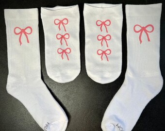 May include: Four white socks with pink bow designs. One sock has a single bow, two socks have three bows, and one sock has a single bow with the word "WOOT" on the bottom.