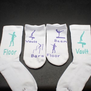 Puede incluir: Cuatro calcetines blancos hasta el tobillo con diferentes eventos de gimnasia impresos en ellos. Los calcetines tienen letras e imágenes moradas y verde azuladas. Los eventos son suelo, salto, barras, viga y suelo.