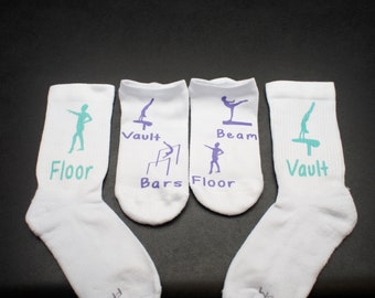 May include: Four white ankle socks with different gymnastics events printed on them. The socks have purple and teal lettering and images. The events are Floor, Vault, Bars, Beam, and Floor.