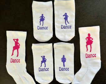 May include: White ankle socks with the word "Dance" and a dancer silhouette in either purple or pink. The socks are arranged on a dark surface.