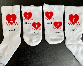 May include: Four white socks with red hearts and different gymnastics events printed on them. The events are Vault, Beam, Floor, and Bars.
