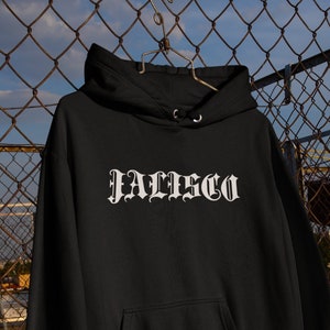Op de afbeelding: Een zwarte hoodie met lange mouwen, een kangoeroezak aan de voorzijde en het woord JALISCO in een wit gotisch lettertype op de borst gedrukt. Het kledingstuk is voorzien van geribbelde boorden aan de mouwen en aan de onderzijde.