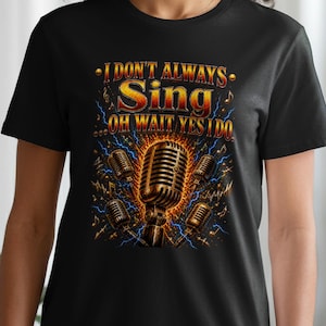 Op de afbeelding: Een zwart t-shirt met korte mouwen en een grafische print van een vintage-stijl microfoon, omringd door kleinere microfoons, blauwe bliksemschichten en muzieknoten. De tekst luidt: I DON'T ALWAYS Sing ...OH WAIT YES I DO.