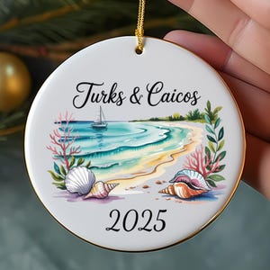 Puede incluir: Adorno navideño redondo de cerámica blanca con borde dorado, que representa una escena de playa con el texto "Turks & Caicos" y "2025". El diseño incluye conchas marinas, un velero y coral, con una cuerda dorada para colgar.