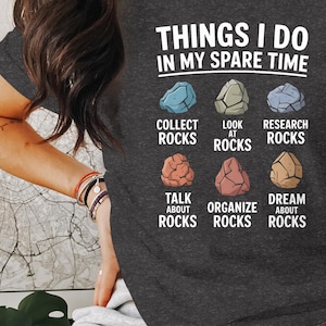 Peut inclure: Un t-shirt gris foncé avec le texte "THINGS I DO IN MY SPARE TIME" et un graphique de six rochers colorés avec du texte décrivant des activités liées aux rochers. Le texte comprend "COLLECT ROCKS", "LOOK AT ROCKS", "RESEARCH ROCKS", "TALK ABOUT ROCKS", "ORGANIZE ROCKS", et "DREAM ABOUT ROCKS".