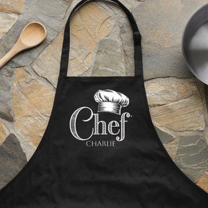 Pode incluir: Avental de cozinha preto com um chapéu de cozinheiro branco e o texto "Chef Charlie" impresso na frente.