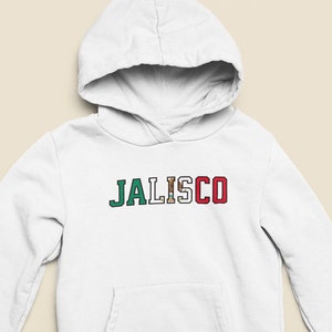 Op de afbeelding: Een wit sweatshirt met capuchon en lange mouwen, voorzien van een kangoeroezak aan de voorzijde en het woord JALISCO op de borst. De letters zijn gekleurd in groen, wit en rood, waarbij de letter I een klein embleemdetail bevat.