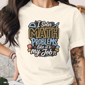 以下が含まれることがあります： 「I Solve MATH PROBLEMS like it's my Job」というテキストと、分度器、コンパス、幾何学模様のイラストが青、黄、黒のインクでプリントされた、クリーム色の半袖Tシャツ。