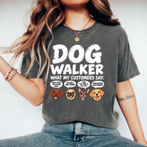 Könnte beinhalten: Ein anthrazitfarbenes Kurzarm-T-Shirt mit dem weißen Text DOG WALKER WHAT MY CUSTOMERS SAY und vier illustrierten Hundeköpfen mit Sprechblasen, die die Sätze TRULY GOES THE EXTRA MILE, RELIABLE AND CARING, I HAD THE TIME OF MY LIFE und PROFESSIONAL AND FRIENDLY enthalten.