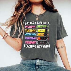 Może przedstawiać: Ciemnoszary t-shirt z napisem BATTERY LIFE OF A TEACHING ASSISTANT oraz pięcioma ikonami baterii oznaczonymi od poniedziałku do piątku. Baterie pokazują malejący poziom naładowania z kolorowymi, wyrazistymi buźkami od radosnych po wyczerpane.