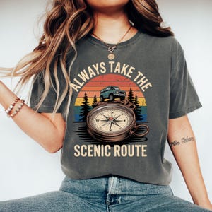May include: Dark gray t-shirt with a graphic featuring a compass, a green SUV, and a sunset design. The text reads "ALWAYS TAKE THE SCENIC ROUTE". The shirt is a comfortable, casual style.