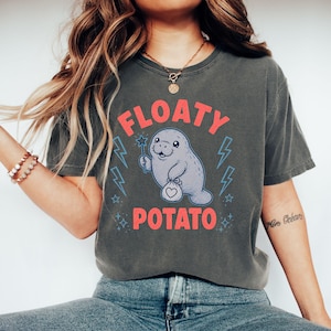 May include: A charcoal gray t-shirt featuring a graphic of a light purple manatee holding a magic wand and a small pouch with a heart icon. The text "FLOATY POTATO" is printed in coral red above and below the illustration, accented by blue lightning bolts and stars.