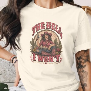 Peut inclure: Un t-shirt à manches courtes de couleur crème avec une impression graphique représentant une cow-girl portant un chapeau et des bottes, tenant un pistolet, entourée de cactus et de fleurs. Le texte "THE HELL I WON'T" est imprimé en lettres rouges.