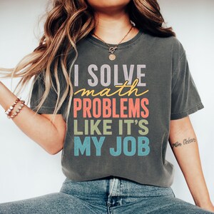 Könnte beinhalten: Ein anthrazitfarbenes Kurzarm-T-Shirt mit dem Text I SOLVE math PROBLEMS LIKE IT'S MY JOB in einem gestapelten, mehrfarbigen Design. Die Wörter sind in den Farben Hellgrau, Gelb, Koralle, Hellgrün und Hellblau aufgedruckt.