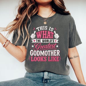 May include: A charcoal grey short-sleeve t-shirt featuring the text THIS IS WHAT THE WORLD'S GREATEST GODMOTHER LOOKS LIKE in pink and white lettering, accompanied by two cartoon hands giving a thumbs-up gesture and small pink heart accents.