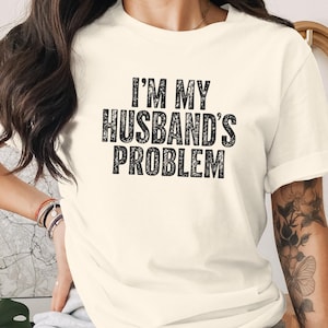 Könnte beinhalten: Ein cremefarbenes Kurzarm-T-Shirt mit dem schwarzen Text "I'M MY HUSBAND'S PROBLEM", der in einer fetten, abgenutzten Schriftart auf der Brust gedruckt ist. Das Kleidungsstück hat einen klassischen Rundhalsausschnitt und eine lockere Passform.