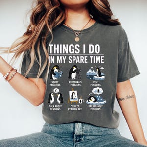 May include: Dark gray t-shirt with the text "THINGS I DO IN MY SPARE TIME" and illustrations of penguins. The shirt features penguins studying, photographing, visiting, talking, collecting art, and dreaming.