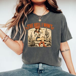 Może przedstawiać: Ciemnoszary t-shirt z krótkim rękawem z nadrukiem graficznym przedstawiającym kowbojkę siedzącą na drewnianym płocie na pustynnym krajobrazie z kaktusami. Napis "THE HELL I WON'T" jest nadrukowany na pomarańczowo nad ilustracją ozdobną czcionką.