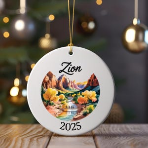 May include: A round white ceramic ornament with a gold string for hanging. The ornament features a landscape scene with the word "Zion" in script above the image and the year "2025" below. The scene depicts a river, bridge, and mountains.