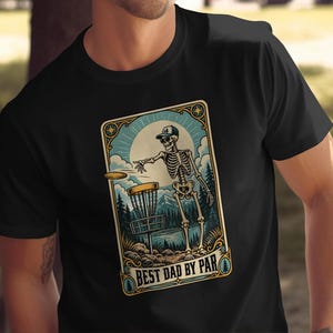 May include: A black t-shirt featuring a graphic of a skeleton wearing a baseball cap and throwing a disc into a disc golf basket. The design includes a mountain landscape, a large sun, and the text "BEST DAD BY PAR" in a decorative frame.