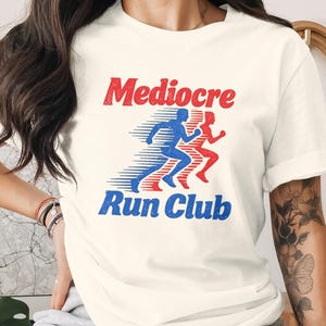May include: A cream-colored short-sleeve t-shirt featuring a graphic print with the text "Mediocre Run Club" in red and blue. The design includes stylized silhouettes of two runners in motion, with blue and red horizontal motion lines.