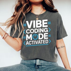 May include: Dark gray t-shirt with the text "VIBE CODING MODE ACTIVATED" in white and blue. The shirt has a relaxed fit and is made of a soft material. The design features a power button graphic.