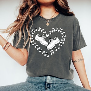 May include: Dark grey t-shirt featuring a white heart design composed of musical notes and two pairs of shoes. The shoes are depicted in a simple, clean style, suggesting a theme related to music or dance.